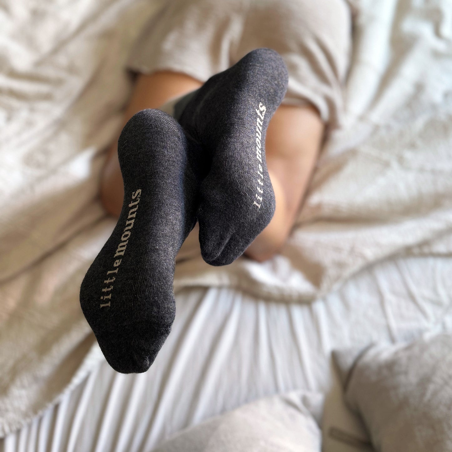 Person wearing dark gray socks with little mounts branding on a light-colored bed.