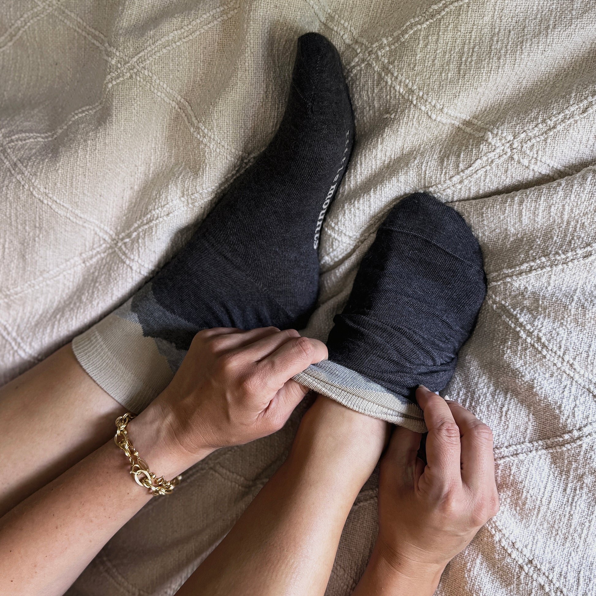 Person putting on dark gray socks with a gold bracelet on a beige textured blanket.